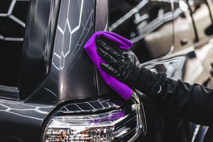 A person cleaning the back of a grey suv with a purple towel.