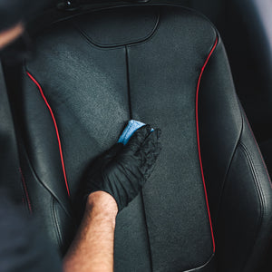 A person wiping a black leather car seat with a blue sponge.