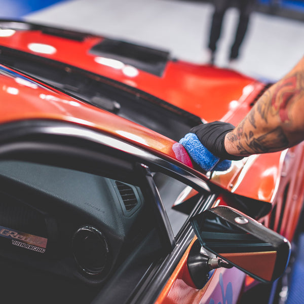 A tatted arm wearing a black rubber glove rubbing an orage Toyota GT86 with Gtechniq Halo Coating with a blue sponge.