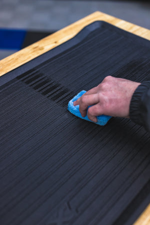 A person scrubbing a car floor mat with a blue sponge.