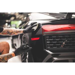 A person scrubbing a black car with one hand and holding a container of Koch Chemie Hand Wax in the other hand. 