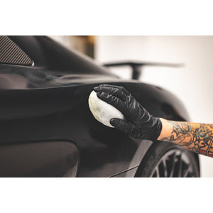 A person scrubbing a black car with a circular white sponge.