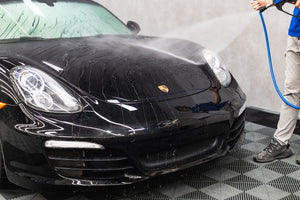 Person washing a black Porsche car with a high-pressure water hose.
