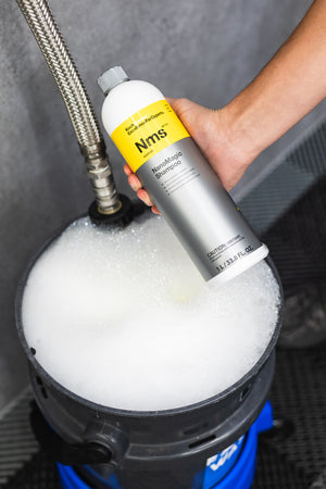 Person holding a bottle of KochChemie NanoMagic Shampoo over a soapy bucket with a pressure washer in the background.