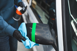 A person spraying KochChemie Panel Preparation Spray onto a blue microfiber to clean a side mirror.