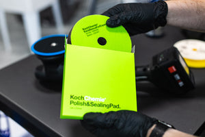 Person holding a Koch Chemie polish and sealing pad with gloves on, against a blurred background.