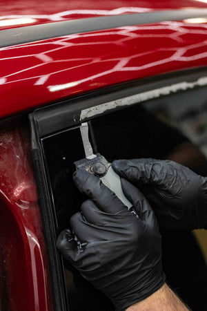 Person using a KochChemie Scraper tool on a car window of a red vehicle.