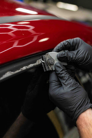 Person using a KochChemie Scraper tool on a car window of a red vehicle.