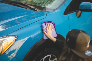 A closeup of a person wearing a hat wiping a blue car with a Minx Royale towel.