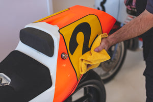 Person cleaning the tail section of a motorcycle with a bright orange and yellow paint job using a yellow microfiber cloth.