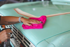 A hand wiping a teal car hood with a pink towel.