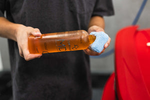 Person holding a bottle of Opti-Coat MDR and a blue sponge near a red vehicle.