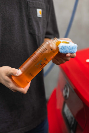 Person holding a bottle of Opti-Coat MDR and a blue sponge near a red car.