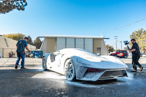 A sports car covered in white foam being washed by a person with a pressure washer, while another person films the process with a camera in an outdoor setting.