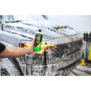 A person holding a bottle of P and S Wide Open in front of a black truck covered in foam and bubbles.