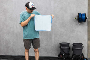 Person holding a blue ftw microfiber towel against a gray wall next to buckets and a hose.