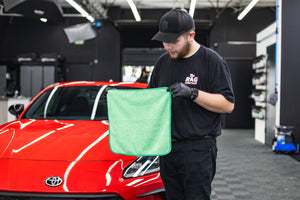 Person holding a green FTW microfiber towel in front of a red car in a garage.