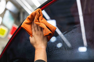 Person cleaning the window of a red car with a orange FTW microfiber towel.