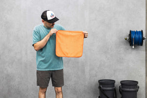 Person holding an orange FTW microfiber towel against a gray wall next to buckets and a hose.