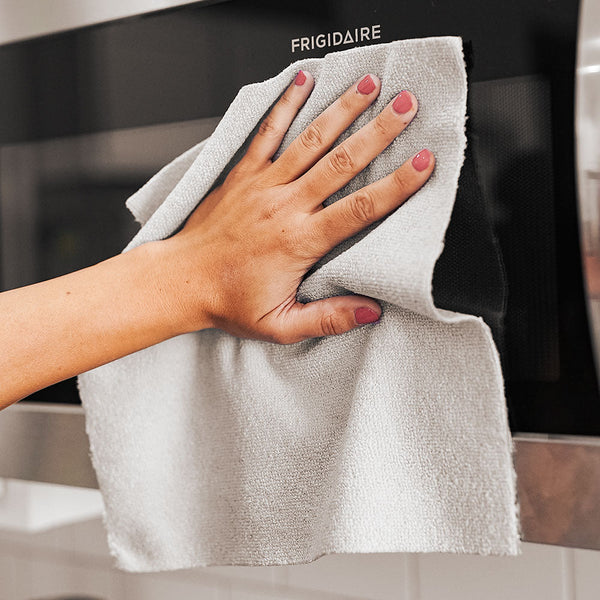 A woman with pink painted fingernails using a single Rip n Rag multi-purpose microfiber towel from The Rag Company in a grey color to clean a microwave oven