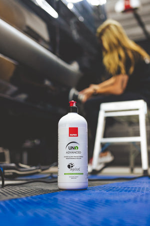 Close-up of a bottle of RUPES UNO Advanced polishing compound on a blue mat, with a person in the background using a polisher on a vehicle.