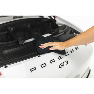 A person scrubbing the back of a car with a black spectrum towel.