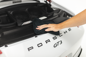 A person scrubbing the back of a car with a black spectrum towel.