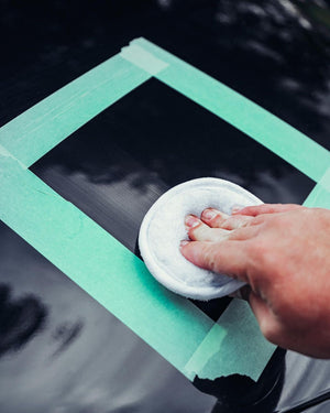 Close-up of a hand using a microfiber applicator pad to polish a black car surface, bordered by green masking tape.