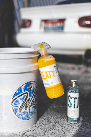 A spray bottle of Glatt Rinse Aid Spray hanging on the side of bucket by its handle with a bottle of Stjarnagloss Gummi under it.