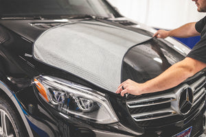 A person displaying The Big One towel over a black car hood.