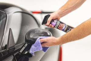 A person spraying a side mirror with P and S Paint Gloss while scrubbing the mirror with a purple Eaglet towel.