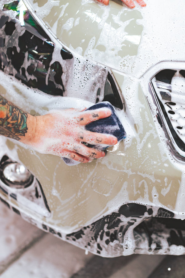 Close-up of a hand washing a car with an Ultra Sponge, both covered in thick suds.