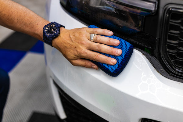 A person is scrubbing the front of a white car with the Ultra Clay Scrubber.