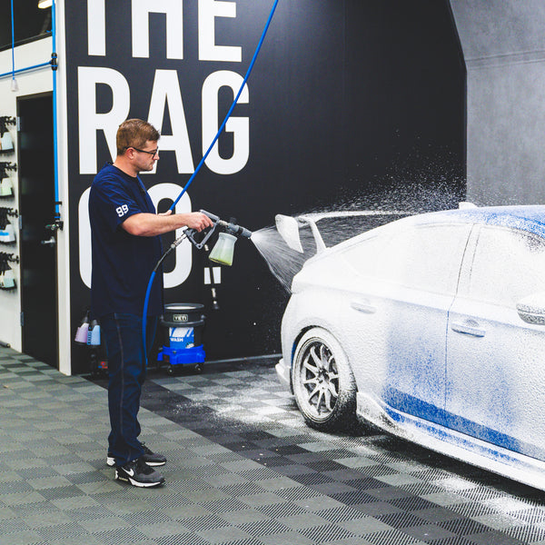 A person using a TRC Ultra H2O Foam Cannon to spray soap onto a blue car.