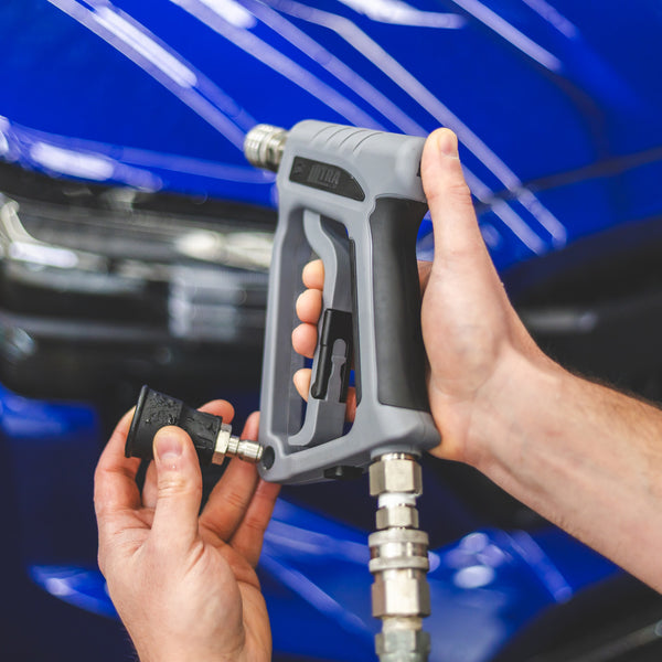 A close up of a person putting a nozzle into the holder on a TRC Ultra H2O Spray Gun with a blurry blue car in the background.