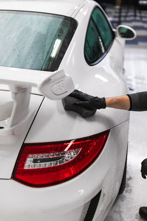 A person wearing black rubber gloves wiping a white Porsche gt3 with the Ultra Safe Sponge. 