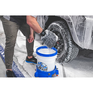 A person rinsing a TRC Ultra Wash Mitt in a wash bucket with a car tire covered with foam in the background.