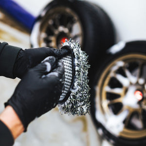 A person wearing black rubber gloves holding an Ultra Wheel Mitt in front of blurred tires.