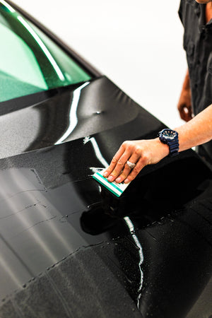 A person using a green squeegee to remove droplets off the hood of a black car.