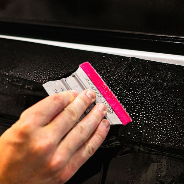 Close-up of a hand using a Yellotools Platinum Squeegee tool with a pink edge to remove water droplets from a car door.