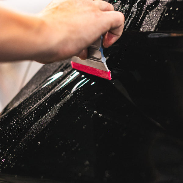 A hand using a Yellotools Platinum Squeegee tool with a pink edge to lay black vinyl onto the outside of a vehicles body.