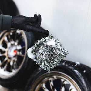 A person dangling a foamy Ultra Wheel Mitt on their index finger with a background of two blurred tires.